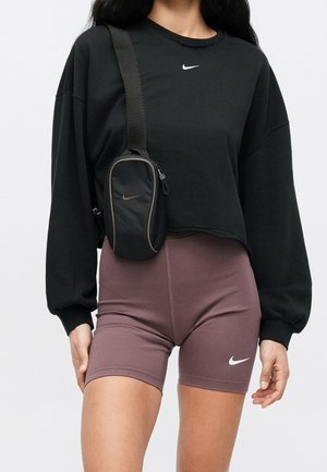Osoba nosząca czarny krótki sweter z długim rękawem oraz fioletowe rowerowe spodenki Nike, z małą czarną torbą na ramię z logo Nike.