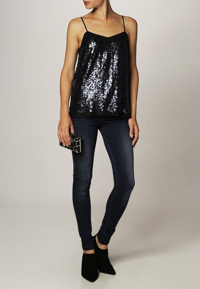 Svart paljett-topp med tunna axelremmar, lös passform, kombinerad med mörka skinny jeans och svarta ankelskor. Håller en mönstrad cluch.