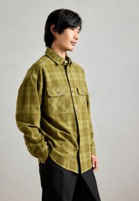 Chemise à carreaux verts avec deux poches poitrine, ourlet arrondi et manches longues. Tissu lisse avec une finition légèrement texturée.