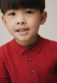 Rood gebreid poloshirt met een gestructureerd patroon, drie bruine knopen en een klassieke kraag. Close-up van het glimlachende gezicht van een kind.