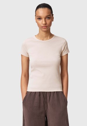 Jeune femme aux cheveux foncés attachés, portant un t-shirt beige à manches courtes et un pantalon marron ample, les mains dans les poches.