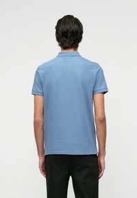 Világoskék pólóing gallérral, rövid ujjal és laza szabással. Simára kidolgozott textúrával és egyszínű dizájnnal, sötét nadrággal párosítva.