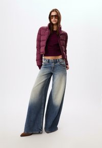 Bordeaux pufferjakke, cropped længde, tætsiddende design. Mørk lilla langærmet top. Bredbenede lyseblå jeans med falmende effekt. Brune hælede støvler.