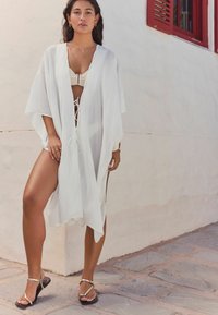 Balts, viegls kimono apvalks ar atvērtu priekšpusi, brīvām piedurknēm, saites aizdari jostasvietā un sānu šķēlumiem. Uzvilkts virs bēša bikinī.