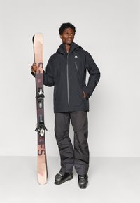 Casaco impermeável preto com bolsos com fecho, combinado com calças de esqui escuras e botas de esqui pretas. Um esqui com prisões é segurado numa das mãos.
