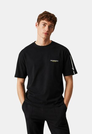 Giovane uomo con capelli castani ricci che indossa una t-shirt nera Trussardi con piccolo logo sul petto e grafica sulla manica sinistra, mani nelle tasche.