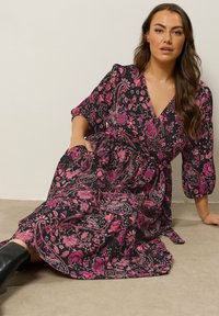 Long black dress with pink floral and paisley patterns, V-neckline, three-quarter sleeves, and a gathered waist, paired with black boots.