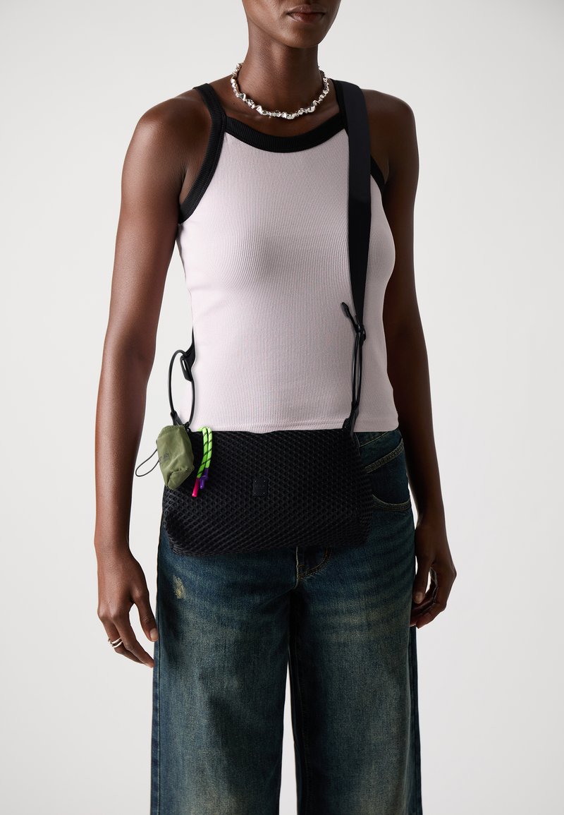 Svart mesh crossbody-veske med justerbar stropp, med en grønn lomme festet med fargerike detaljer. Båret over en lys tanktop og jeans.