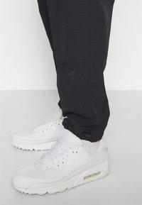 Bílé tenisky Nike Air Max se síťovinovými a koženými panely, doplněné černými texturovanými kalhotami s šňůrkou u kotníku.