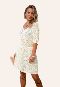 Gebreide jurk in crème met zigzagpatroon, V-hals, driekwartmouwen en een taille met trekkoord. G gedragen met een geweven tas en gelaagde sieraden.
