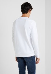 Sudadera blanca con escote redondo y mangas largas. Presenta puños y dobladillo de canalé, con textura suave. Llevada con jeans azules, vista desde atrás.