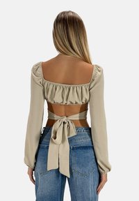 Top beige a maniche lunghe con scollo a balze e legato sul retro. Il tessuto sembra morbido; abbinato a jeans blu a vita alta in una vestibilità rilassata.