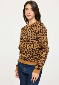 Pull en marron avec imprimé léopard noir, poignets et ourlet côtelés. Texture douce avec un design classique à col rond. Porté avec un jean bleu.