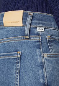 Ljusblåa denimjeans med en läderbakficka märkta "CITIZENS of HUMANITY." Inkluderar gul söm och en liten vit etikett.