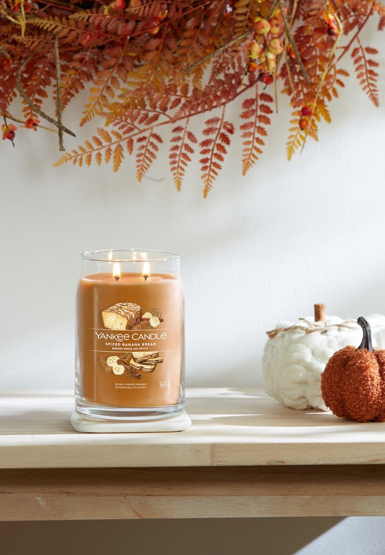 Yankee Candle in einem klaren Glasbehälter, orangefarbener Wachs, beschriftet "Gewürzter Bananenbrot", umgeben von dekorativen Kürbissen auf einem Holzregal.
