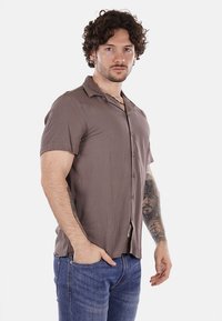 Camicetta a maniche corte con bottoni in tessuto marrone tenue, caratterizzata da una superficie liscia e un leggero lucido. Indossata con jeans blu e tatuaggi visibili.