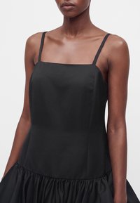 Femme portant une robe noire sans manches avec de fines bretelles et un détail de jupe froncée, montrée du cou jusqu'à mi-cuisse sur un fond uni.