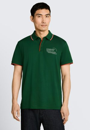 Mężczyzna w ciemnozielonej koszulce polo z czerwonym wykończeniem i napisem "Forever Summer Tropical Paradise" na piersi, stojący na jasnym tle.