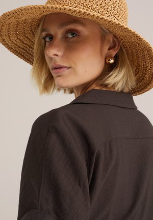 Vrouw met kort blond haar die een strohoed met brede rand, gouden ronde oorbellen en een donkerbruin overhemd met kraag draagt, kijkt over haar schouder.