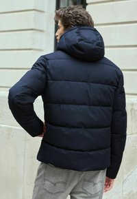Jachetă puffer de marine cu glugă, dotată cu secțiuni cusute pentru izolație, fabricată dintr-un material durabil, asociată cu blugi gri deschis.