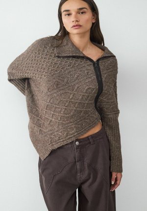Brun strikket pullover med en tekstureret kabel-design, høj krave og en sidelukning, parret med mørkebrune brede bukser.