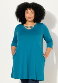 Tunica teal realizzata in tessuto morbido, con scollo a V e design incrociato, maniche a tre quarti e tasche laterali. La forma svasata offre comfort.