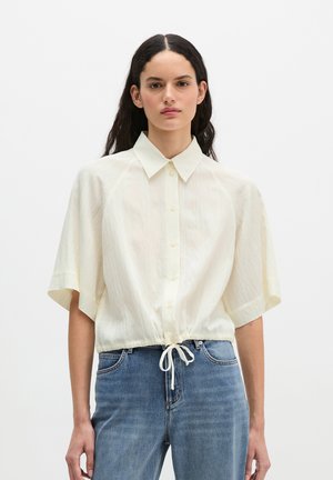 Vrouw met lang donker haar draagt een crèmekleurig oversized kortermig overhemd met knopen en een trekkoord in de taille en een blauwe spijkerbroek, staat rechtop.