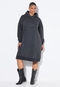 Dunkelgraues Kapuzenkleid aus weichem Stoff, mit langen Ärmeln, lockerem Schnitt und abgerundetem Saum. Kombiniert mit schwarzen Cowboy-Stiefeln.