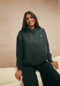 Donkergrijze hoodie met een voorzak en geborduurd "self." logo. Losse pasvorm met een gestructureerde stof. Gecombineerd met zwarte broek.