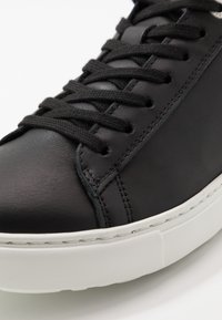Sneaker en cuir noir avec des lacets noirs, une texture lisse et une semelle en caoutchouc blanc contrastant avec des détails de couture visibles.