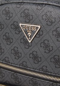 Zaino in tessuto nero con texture, caratterizzato da un prominente logo triangolare argento "GUESS" e dettagli in zip dorati. Accenti a motivi sottili.