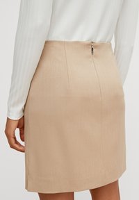 Jupe beige tailleur arrivant au genou, avec texture verticale, fente latérale et fermeture éclair au dos. Associée à un top blanc côtelé à manches longues.
