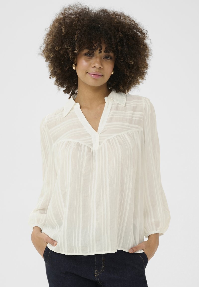 Blusa a righe bianche realizzata in tessuto leggero. Presenta un collo a V, una chiusura con bottoni e maniche lunghe con polsini elastici.