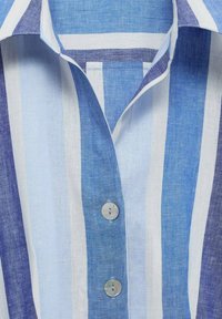 Cămașă din in blue și alb cu dungi, având un guler clasic și nasturi din scoică naturală. Dungile variază ca lățime și nuanță de albastru.