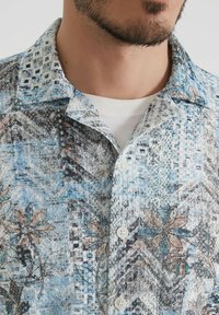 Homme portant une chemise à boutons texturée avec des motifs abstraits bleus, gris et bruns sur un t-shirt à col rond blanc.