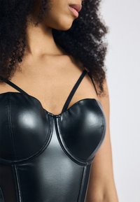 Zwarte faux leren korsett met gevormde cups, dunne bandjes en een gladde, glanzende afwerking. Kenmerkt zichtbare stiksels en een getailleerd ontwerp.