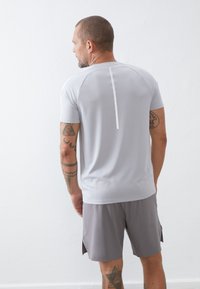 Graues sportliches Shirt mit kurzen Ärmeln und einem reflektierenden Streifen, kombiniert mit grauen Shorts, von hinten betrachtet. Glattes, leichtes Material.