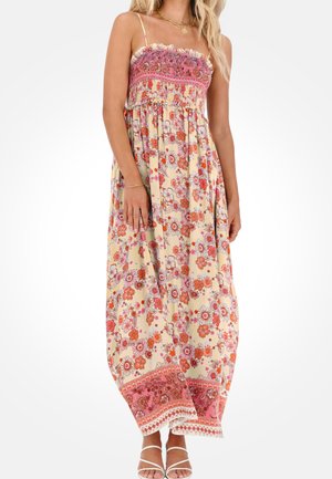 Vrouw draagt een bloemenpatroon strapless maxi-jurk met een roze gerimpelde top en witte sandaaltjes met bandjes, staande tegen een effen achtergrond.