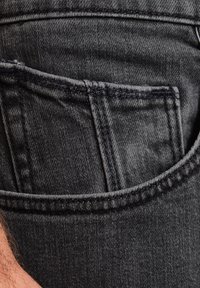 Detail van de zak van een zwarte denim broek, met een gestructureerde stof, opvallende stiksels en een zichtbaar klein zakje.