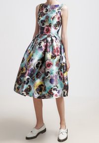 Vestido con estampado floral que presenta un diseño sin mangas, un torso ajustado y una falda acampanada en verde azulado con patrones de pensamientos en púrpura y rosa. Zapatos blancos.