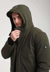 Giacca isolante verde oliva con cappuccio, dotata di zip frontale e tasche laterali. Il materiale ha una texture liscia; include il logo sulla manica.
