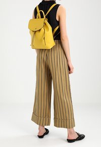 Sac à dos en cuir jaune avec rabat supérieur, fermeture à cordon et bretelles réglables. Associé à un pantalon large à rayures beige.