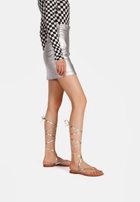 Nainen, jolla on ykkösmallinen mustavalkoinen ruudullinen paita, hopeanvärinen metallinen mini-hame ja kultaiset gladiatorisaappaat, jotka on sidottu pohkeisiin, seisoo valkoista taustaa vasten.