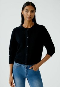 Gilet noir avec un col rond et des manches longues, comportant des boutons et un tissu doux et texturé. Porté par-dessus un jean bleu.