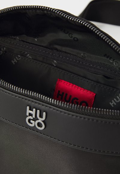 Sac en nylon noir avec une texture lisse, doté d'une fermeture éclair visible et d'un logo "HUGO" en argent. À l'intérieur, une étiquette rouge avec le branding "HUGO".