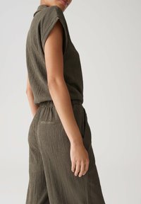 Olive green jumpsuit with a textured fabric, short sleeves, and an elastic waistband. Features side pockets and a relaxed fit.