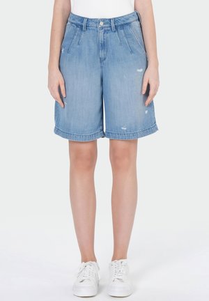Personne portant un short en denim bleu clair jusqu'aux genoux avec une légère usure et des baskets blanches, se tenant devant un fond uni.