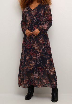 Frau, die ein langes, dunkles Kleid mit Blumenmuster und langen Ärmeln sowie schwarzen Stiefeletten trägt, steht vor einem einfarbigen hellen Hintergrund.