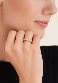 Anello d'oro con una fascia texturizzata e una pietra preziosa rettangolare e trasparente. L'anello è indossato su un dito, mostrando il suo design su uno sfondo neutro.