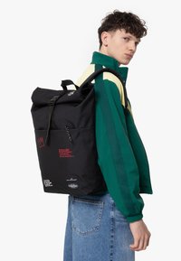 Svart rulltoppsryggsäck med framficka, röda textdetaljer och ett robust bälte. Bärs av en person i en grön och gul jacka.